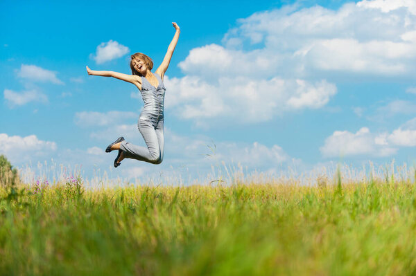 Young beautiful woman in a blue denim overalls fun jumps up against the blue sky with clouds and green grass year high.