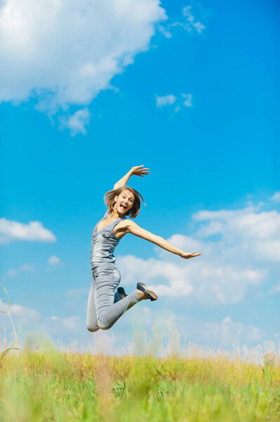 Young beautiful woman in a blue denim overalls fun jumps up against the blue sky with clouds and green grass year high.