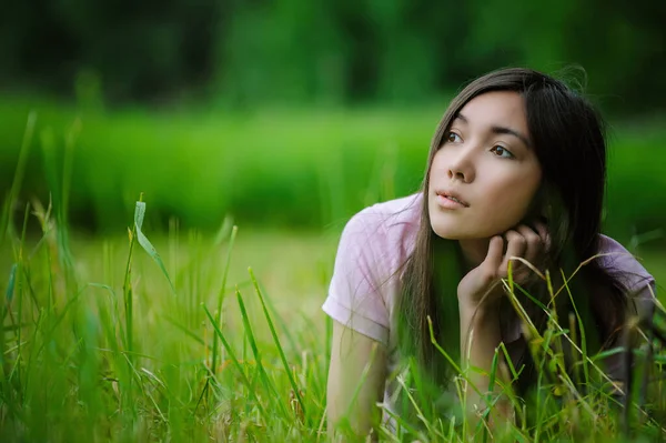 身穿粉色衬衫和蓝色牛仔裤的年轻而沉思的美丽的亚洲女人躺在草地上 旁边是一个绿色的夏季公园 — 图库照片