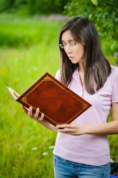 身穿粉色衬衫和眼镜的年轻而沉着的美丽的亚洲女人在一个绿色的夏季公园的背景下看了一本红皮相册 — 图库照片