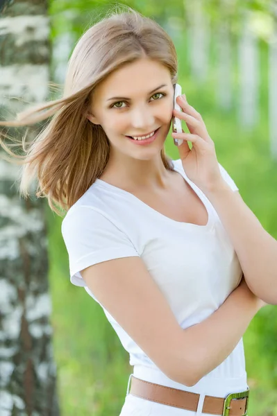 夏の緑の都市公園を背景に携帯電話で話している白いブラウスの若いかなり笑顔の女性 — ストック写真