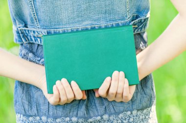 Kot ceketli genç kadın yeşil bir şehir parkının elinde bir kitap tutuyor..