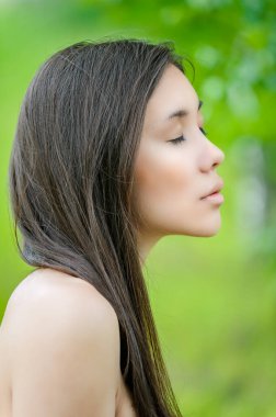 Genç, sakin, uzun saçlı, gözleri kapalı ve arka planda çıplak omuzları olan Asyalı bir kadın. Yeşil şehir parkında..