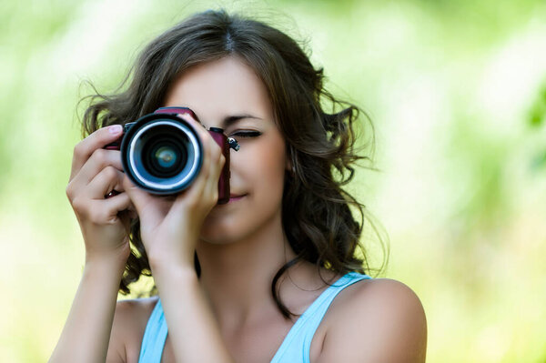 Молодая красивая спокойная женщина в голубой футболке снимает на камеру на фоне летнего зеленого городского парка.