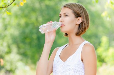 Beyaz bluzlu, çekici, sakin bir kadın yaz mevsiminde yeşil bir parkta bir şişeden su içiyor..