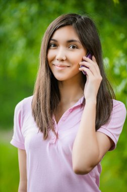 Yeşil şehir parkında güneşli bir yaz gününde akıllı telefonla konuşan mutlu Asyalı bir kadın..