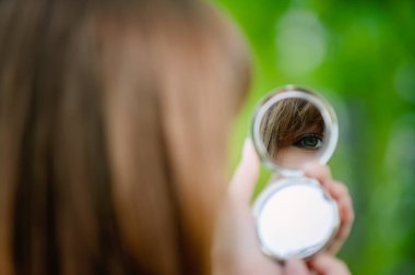 Genç ve sakin bir kadın yuvarlak bir aynaya bakar. Yeşil bir şehir parkının arka planına bakar..