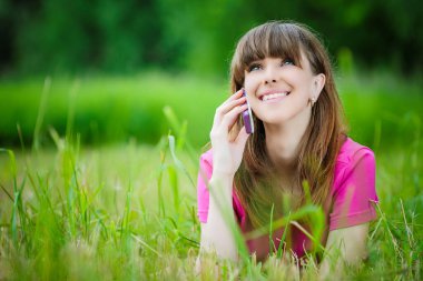 Kırmızı bluzlu, gülümseyen genç bir kadın çimenlere uzanıyor ve yeşil bir yaz parkının arka planında cep telefonuyla konuşuyor..