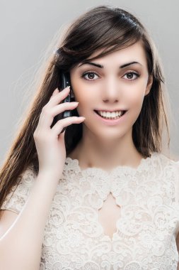 Gri bir arka planda cep telefonuyla konuşan uzun siyah saçlı, gülen güzel bir kadın..