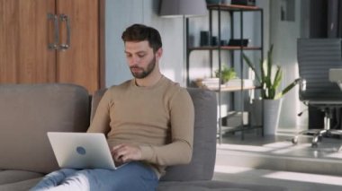 Smiling man typing freelancer with laptop on couch at home.