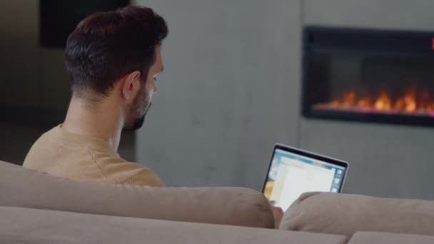 Молодой человек работает с ноутбуком на диване у камина дома. Мужчина с ноутбуком на диване у камина — стоковое видео