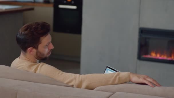 Denkende man werkt met laptop op de bank bij de open haard thuis. Een man met een laptop op de bank bij de open haard — Stockvideo