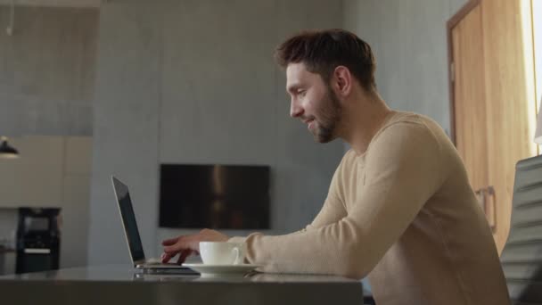 Νεαρός χαρούμενος άντρας πληκτρολογεί στο λάπτοπ από το γραφείο του σπιτιού. Ελκυστικό μήνυμα δακτυλογράφησης ανθρώπου σε φίλους — Αρχείο Βίντεο