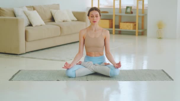 Entspannte junge Frau in Sportbekleidung meditiert in Lotusposition sitzend auf Yogamatte mit geschlossenen Augen — Stockvideo