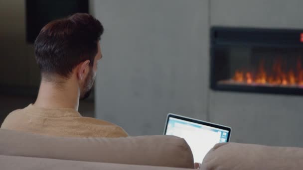 Young profesisonal man working with laptop on the couch by the fireplace at home. A man with a laptop on the couch by the fireplace — Stock Video
