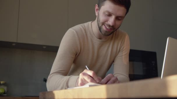 Усміхнений молодий чоловік спілкується відео в прямому ефірі за допомогою комп'ютера. Молодий чоловік робить нотатки — стокове відео