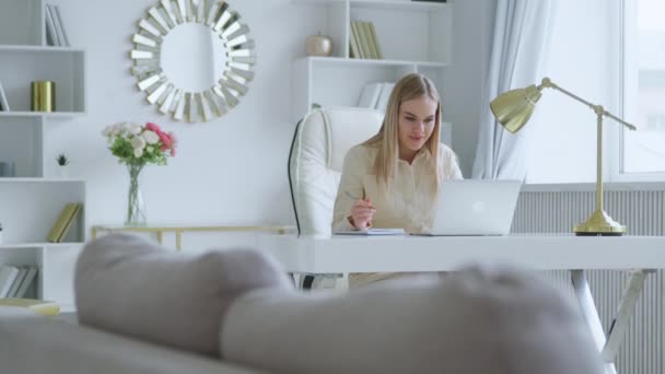 젊은 여성 이 노트북을 이용 한 비디오 콘퍼런스에서 의사소통을 하고 있습니다. 매력적 인 여자가 책상에 있는 공책에 필기를 한다 — 비디오