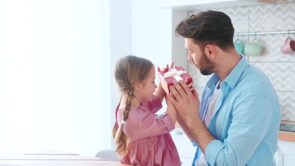 Ung forælder giver en gave til sin lille datter i et hjem interiør. Smilende pige med sin far åbner en fødselsdagsgave. – Stock-video