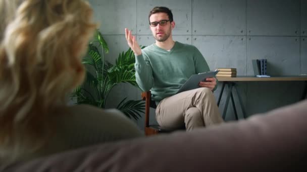 Een jonge professionele psychotherapeut die aan een patiënt uitlegt wat de oorzaken van depressie zijn. Jongeman met klembord bij psychotherapie — Stockvideo