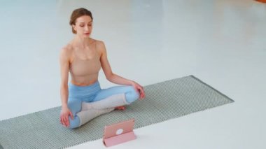 Spor kıyafetli rahat genç bir kadın Lotus pozisyonunda meditasyon yapıyor yoga minderinde gözleri kapalı oturuyor.