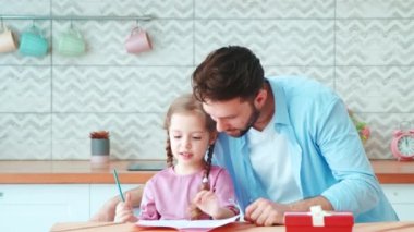 Küçük kızı olan genç bir baba ev ödevi yapıyor. Babası küçük kızına mutfak masasında yazı yazmayı öğretiyor.