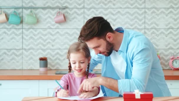 Un padre joven con una hija pequeña haciendo la tarea. Padre enseñando a su hija a escribir en la mesa de la cocina — Vídeo de stock