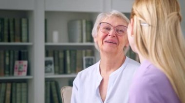 Gözlüklü yaşlı bir kadın kitaplığın arka planında genç bir kıza hediye veriyor.