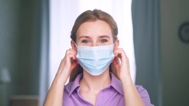 Jovem mulher tirando a máscara azul médica em casa. Menina sorridente sem máscara médica olhando para a câmera — Vídeo de Stock