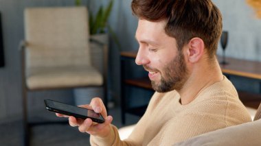 Kanepede oturan adam telefonu evde tutuyor. Akıllı telefonlu gülümseyen genç adam.