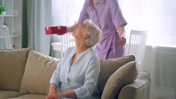 Blonde aux cheveux longs en robe violette donne petite boîte avec cadeau à dame âgée avec sourire joyeux. Fête des mères — Video