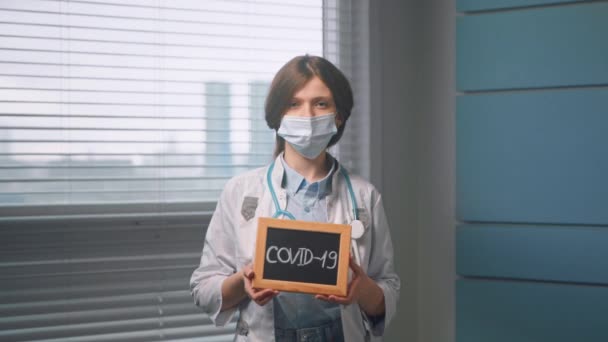 Jonge vrouw arts in het gezicht masker houdt covid 19 naamplaatje — Stockvideo