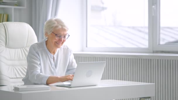 Sonriente señora de edad con el pelo gris y tipos de gafas en el ordenador portátil — Vídeo de stock