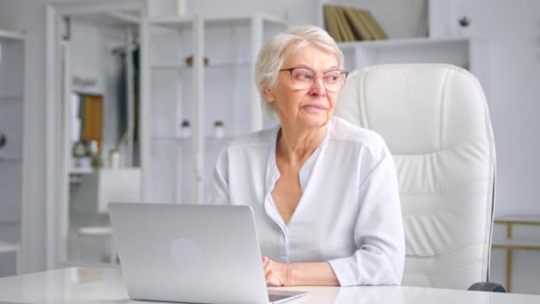 Senior woman with grey hair and glasses glances into laptop — Αρχείο Βίντεο