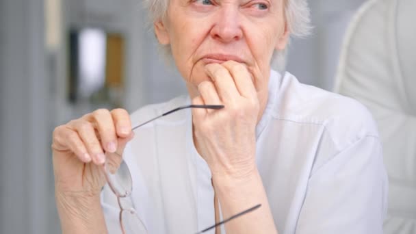 Thoughtful senior woman holds glasses in hand by chin — Stockvideo