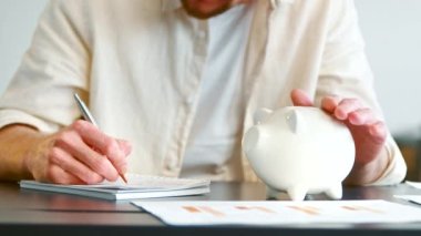 Male person in yellow writes in pad stroking piggy bank