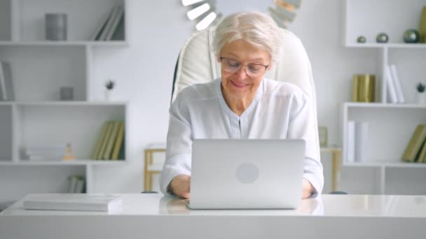 Smiling aged woman in glasses looks at laptop and types — Video
