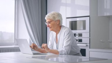 Old woman company manager types on grey laptop in kitchen