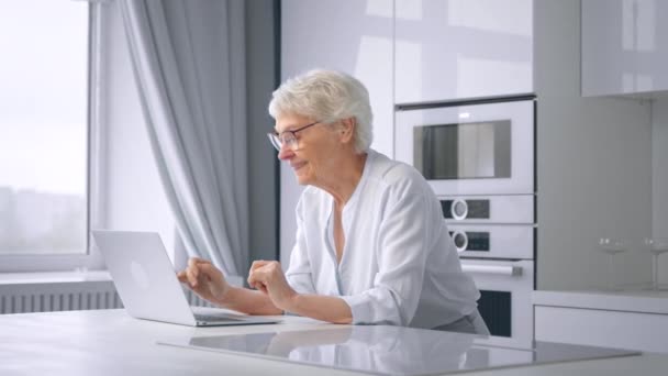 Old woman company manager types on grey laptop in kitchen — Stock Video