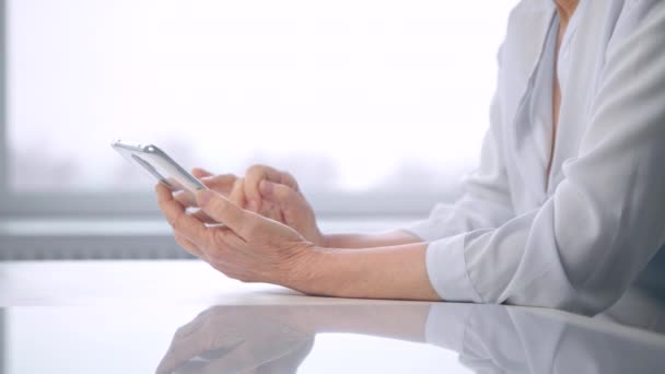 Old woman in blouse wrinkly hands type on white smartphone — Stock Video