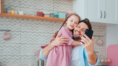 Gülümseyen baba ve kız mutfaktaki akıllı telefondan selfie çekiyorlar.
