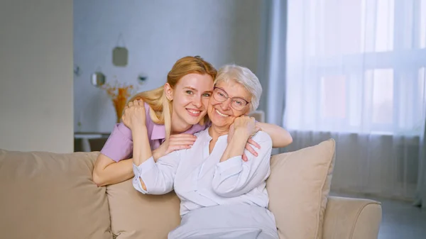 Niña y anciana abrazándose en un sofá. Familia sonriente mirando a la cámara con una sonrisa —  Fotos de Stock