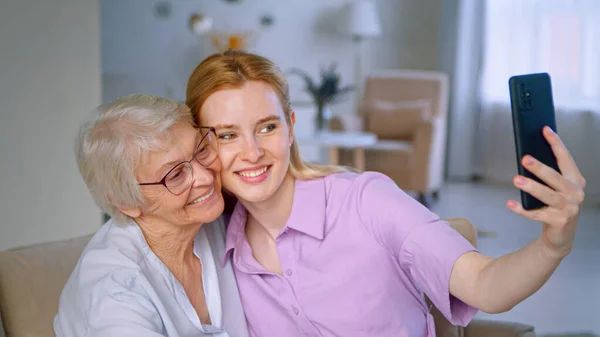 Hija pequeña enseña a la madre anciana a tomar selfie en el teléfono inteligente en casa. Mujeres sonrientes con smartphone — Foto de Stock