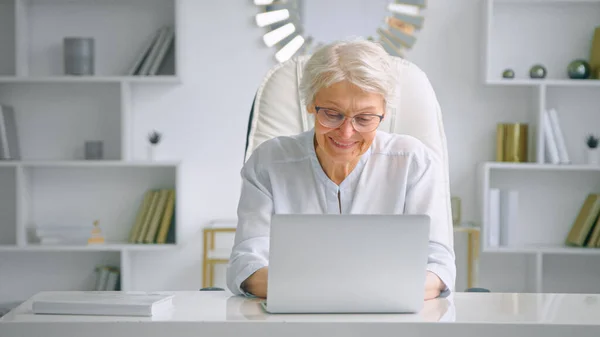 Secretária de senhora sênior sorridente com cabelos grisalhos e óculos em tipos de terno elegante no laptop à mesa — Fotografia de Stock