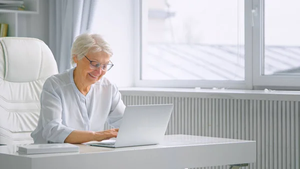 Senhora idosa sorridente com cabelos grisalhos e tipos de óculos no laptop sentado na mesa branca — Fotografia de Stock