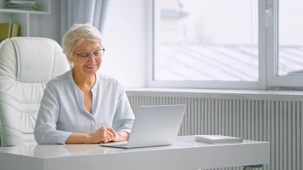 Old company woman worker waves hand greeting colleagues at online meeting and sitting at white table — Stock Photo, Image