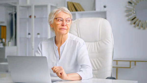 Konzentrierte betagte Managerin in Brille sitzt auf großem Stuhl am Tisch — Stockfoto