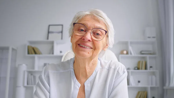 Smiling grey haired aged mother in glasses waves hand greeting adult children and talks at online video — Stock Photo, Image