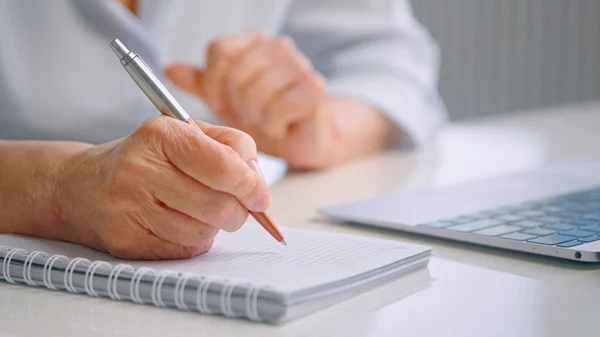 Senior dam lärare med rynkiga händer håller färgad penna över vit sida av papper anteckningsbok nära grå laptop på bordet — Stockfoto
