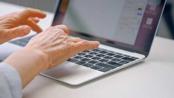 Velha mulher mãos digite no teclado preto do laptop cinza na mesa branca com reflexos de luz brilhante no escritório no trabalho — Fotografia de Stock