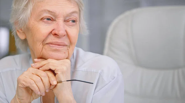 Mujer envejecida con pelo gris corto mira por la ventana sosteniendo gafas en la mano y sonriendo alegremente en la oficina con estilo —  Fotos de Stock